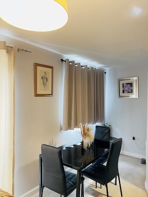 A dining area with a glass table, black chairs, a vase with dried plants, and two framed pictures on the wall.