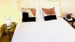 En pænt redt seng med hvide sengetæpper og to pyldepuder, et sengebord med en lampe samt billedkunst over sengen.
