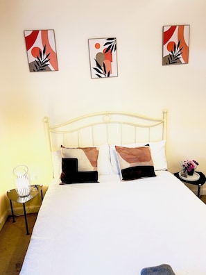 A neatly made bed with white bedding and two decorative pillows, a bedside table with a lamp, and wall art above the bed.