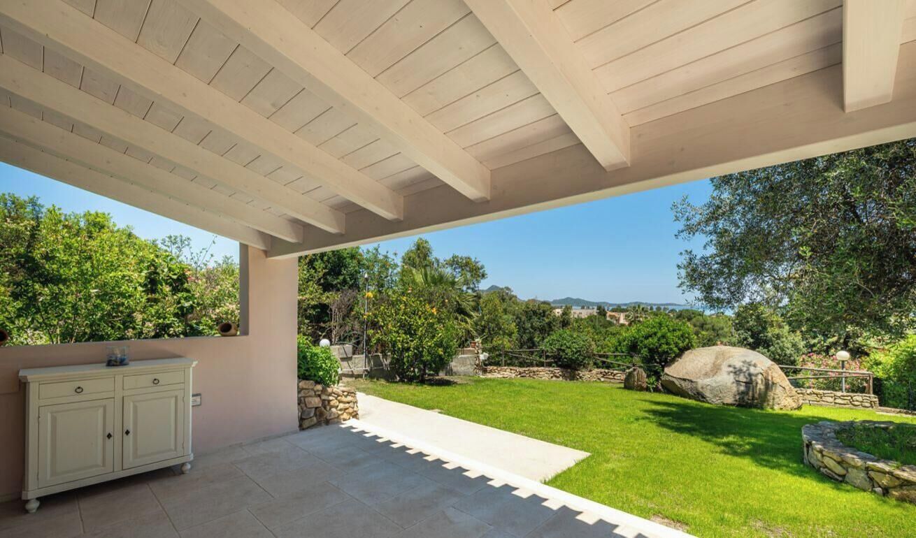 A covered patio area with a stone bench, a cabinet, and a view of a landscaped garden with a large rock and trees.