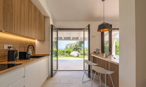 A modern kitchen with an open door leading to a balcony, a coffee machine, and a pendant light. - Villa Reya - Costa Rei Family  Retreat (Costa Rei)