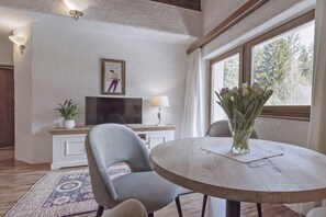 Ein Zimmer mit einem runden Holztisch, zwei grauen Stühlen, einem Fernseher auf einer weißen Kommode, einer Topfpflanze und einer Vase mit Blumen.