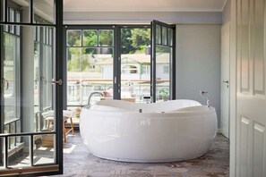 Ein modernes Badezimmer mit einer großen Badewanne, einer Glastür, die auf einen Balkon führt, und Blick auf eine Wohngegend.