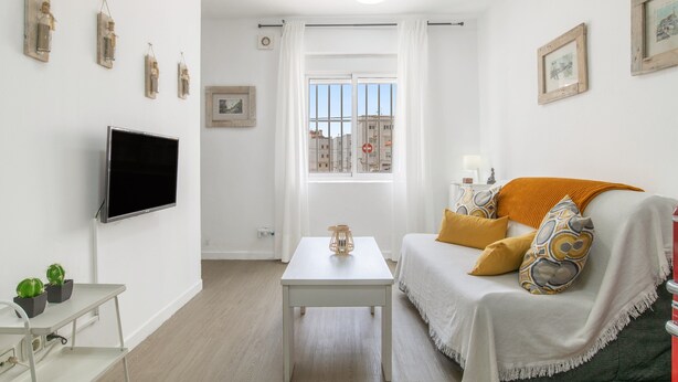 Een woonkamer met een flatscreen televisie, een witte tafel, een bank met opvallende kussens en een raam met uitzicht op gebouwen.