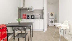 Een kleine keuken met een magnetron, koffiezetapparaat en wasmachine. Er is een eettafel met stoelen en een rode apparatuur.