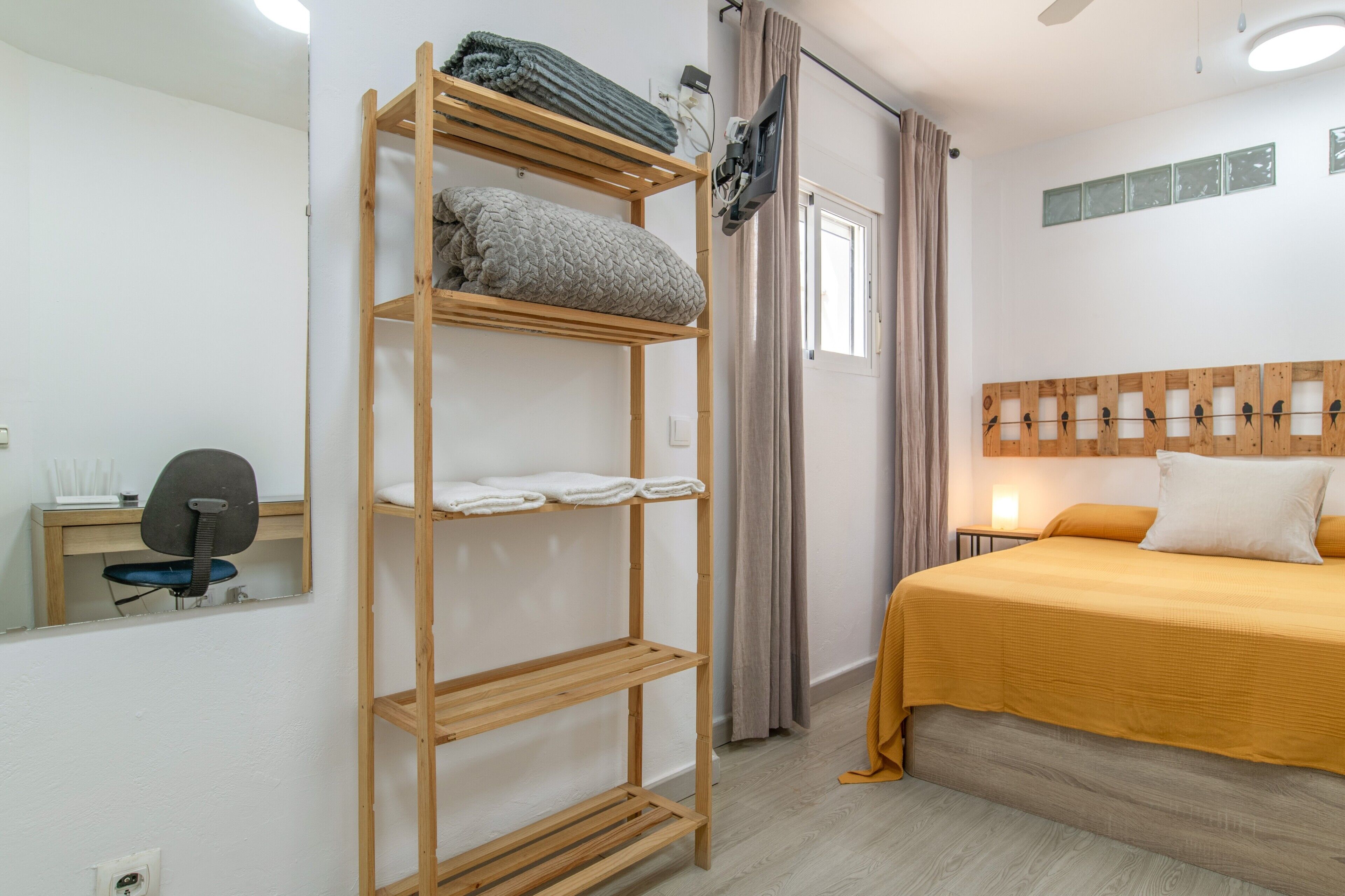 Une chambre à coucher comprenant un lit, une étagère en bois, un bureau avec une chaise, un miroir et une fenêtre avec des rideaux.