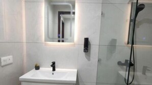 Une salle de bain moderne avec un meuble-lavabo blanc, un miroir, une cabine de douche en verre et une petite poubelle.