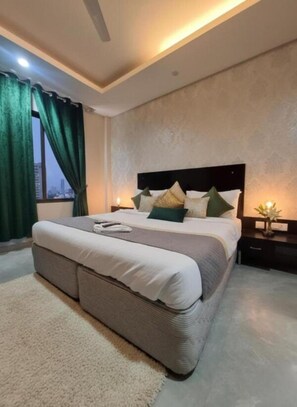 A modern bedroom with a large bed, a nightstand, a bedside table, and a view of the city through a window with green curtains. - Giri Prime Inn Cyber City By OTABoostup (Gurugram)