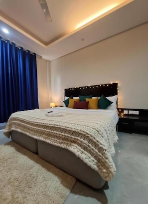 A modern bedroom with a large bed, a headboard with lights, a bedside table, and a blue curtain. - Giri Prime Inn Cyber City By OTABoostup (Gurugram)
