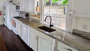 A kitchen with white cabinets, a granite countertop, a sink, and a window with a view of greenery. - Historic Creole Cottage In The Marigny (New Orleans)