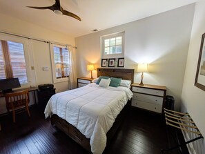 Ein Schlafzimmer mit einem Bett, zwei Nachttischen, einem Schreibtisch, einem Stuhl, einem Deckenventilator und einem Fenster mit Jalousien.