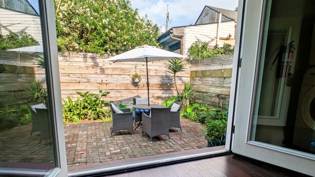 An open sliding glass door leading to a patio with a table and chairs, surrounded by greenery and a brick wall. - Historic Creole Cottage In The Marigny (New Orleans)