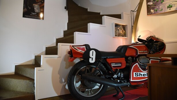 Una motocicleta estacionada en una escalera con un cartel con el número 9, una fotografía en un marco en la pared y un cuadro sobre la escalera.