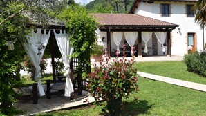 Un área de estar al aire libre con techo, una pérgola, un edificio blanco con techo de tejas y un césped cuidado.