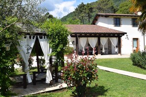 Ein überdachter Außenbereich mit Pergola, ein weißes Gebäude mit Ziegeldach und ein gepflegter Rasen.