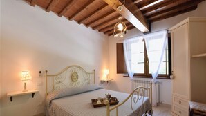 Un dormitorio con cama, lámparas de noche, una ventana con cortinas, un radiador y techo de madera.