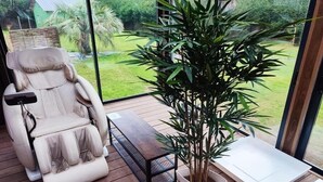 Een massagestoel, een houten tafel, een potplant en een whiteboard op een terras met uitzicht op een tuin.