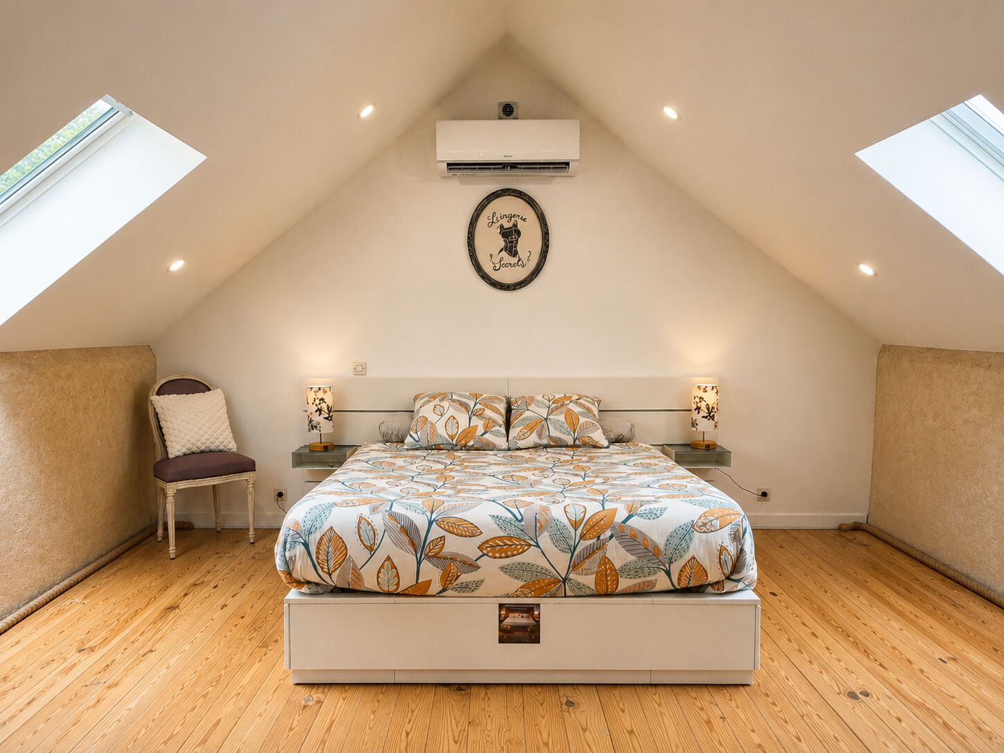A bedroom with a sloped ceiling, a bed with a leaf-patterned bedspread, two bedside lamps, a chair, and an air conditioning unit.