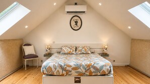 Een slaapkamer met een schuine balkenlaag, een bed met een sprei met bladmotief, twee bedlampen, een stoel en een airconditioning.