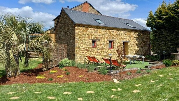 Een stenen huis met schuin dak, een klein portaaltje en een tuin met gras en planten.