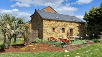 A stone house with a sloped roof, a small porch, and a garden with grass and plants.