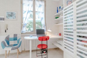 Ein Zimmer mit Schreibtisch, Laptop, Stuhl, einem roten Hocker und einem Regal mit Aufbewahrungsboxen.