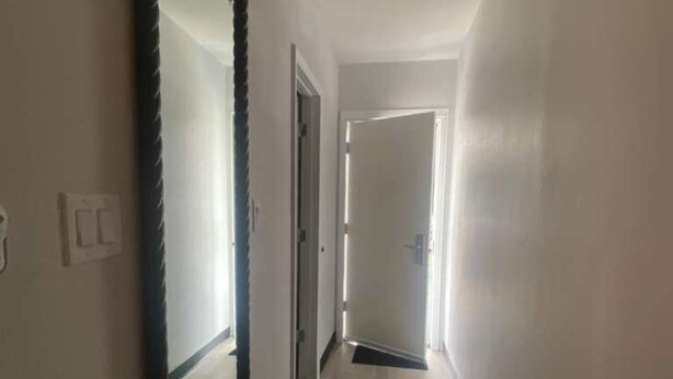 Um corredor estreito com um espelho grande à esquerda, uma porta no final e um tapete de entrada no chão.