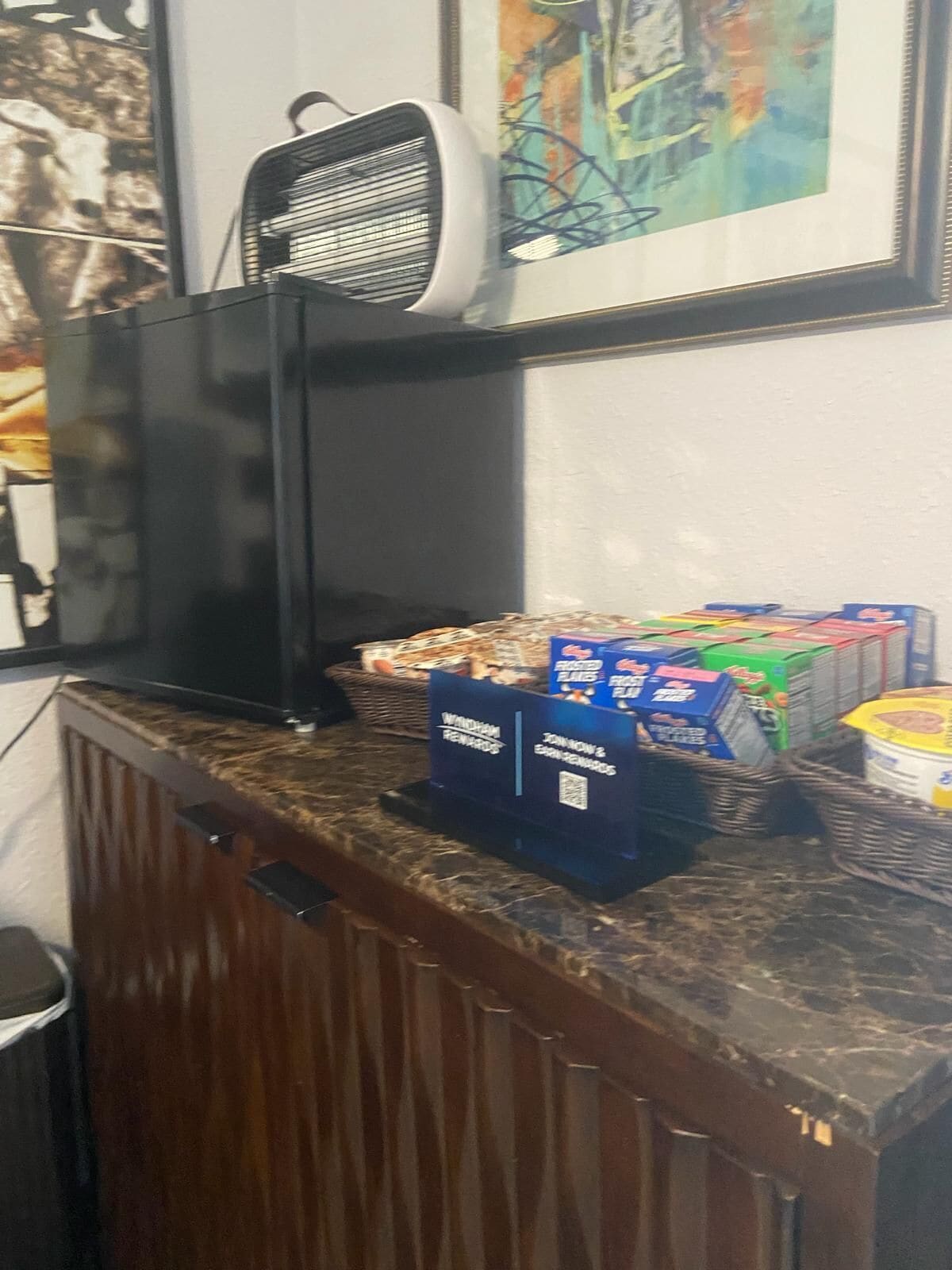 Balcão de recepção com um ventilador, uma pintura abstrata emoldurada e um balcão com caixas de lanches e um recipiente.