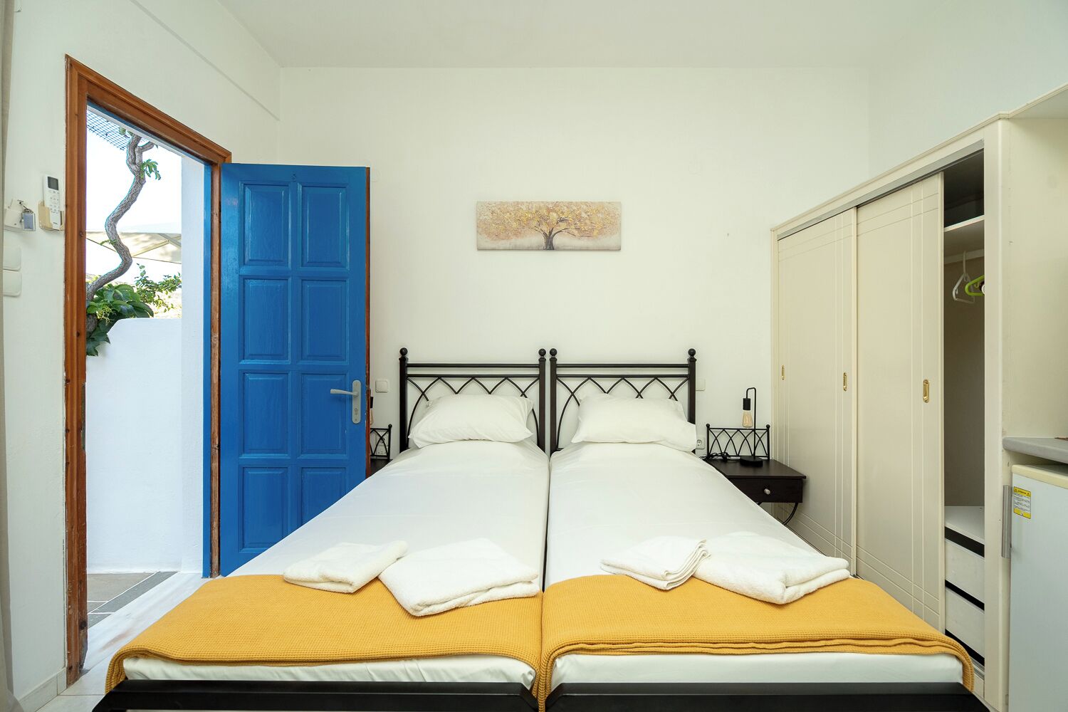 A double bed with a yellow blanket, a blue door, and a white wall with a painting.