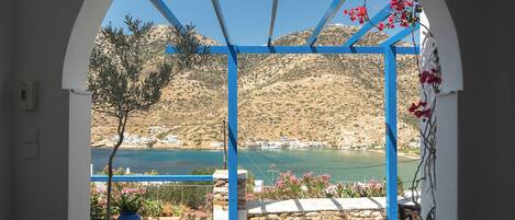 A view through an arched doorway with a blue structure, overlooking a coastal landscape with hills and a body of water.