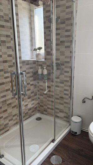 Eine gläserne Dusche mit gefliestem Boden, einer Handstange und einem kleinen Ablagebereich für Toilettenartikel.