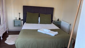 Cama bem arrumada com colcha verde e duas almofadas, manta branca, mesa de cabeceira pequena com abajur e radiador.