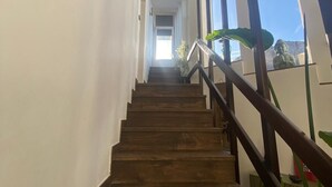 Un escalier en bois, doté d’une rampe, mène à une pièce lumineuse avec une grande fenêtre et une statue.
