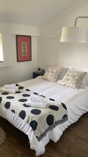 Cama bem arrumada com um cobertor de poá, duas almofadas estampadas com folhas, um abajur e um quadro na parede.