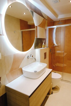 Ein modernes Badezimmer mit einem großen, ovalen Spiegel, einem weißen Waschbecken, einer Duschkabine aus Glas und einer Toilette.