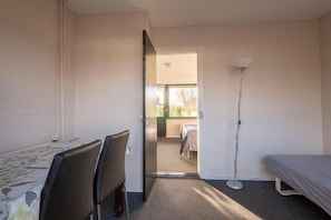 A room with a bed, a desk with a chair, a standing lamp, and a door leading to another room.
