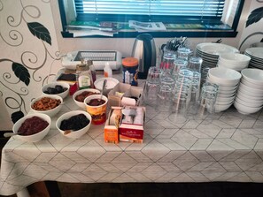 Een tafel met verschillende etenswaren en keukengerei, een raam met jaloezieën, en een betegelde vloer.