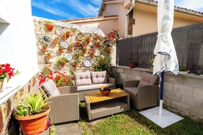 Un patio con mobiliario de mimbre, una pared de piedra con plantas en macetas y un paraguas blanco.