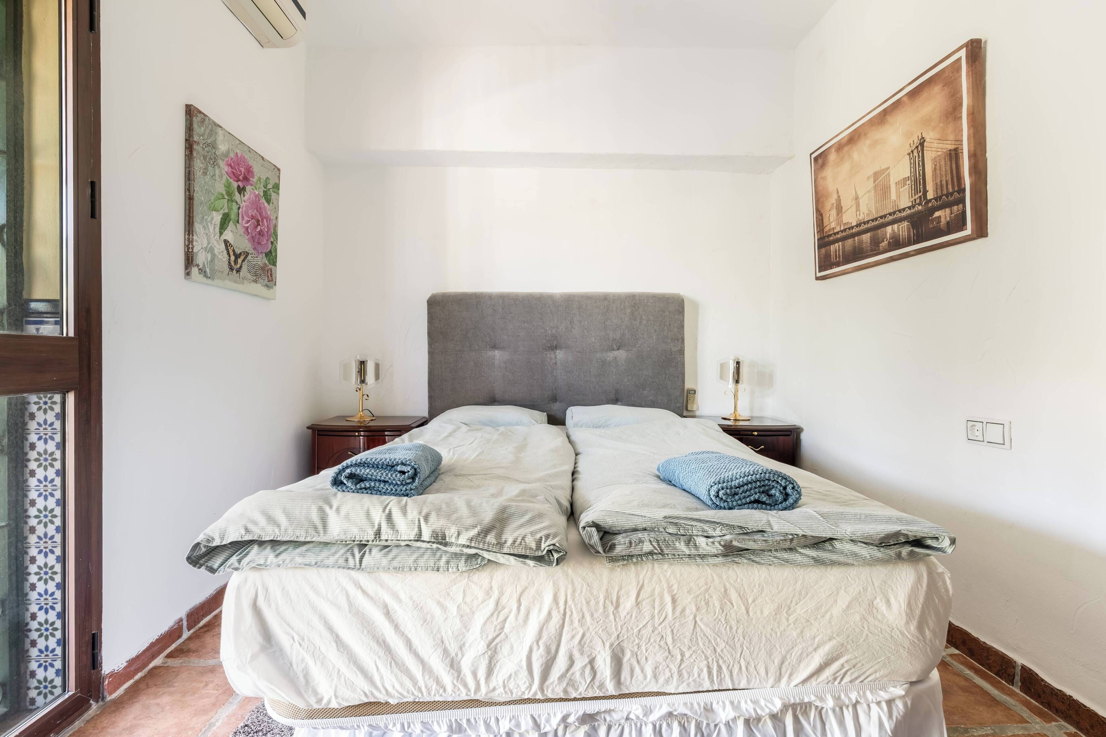 Ein Doppelbett mit zwei Kissen und zwei gefalteten Handtüchern, ein grauer möbelbezogener Kopfseitenverkleidung, ein kleiner Nachttisch mit Lampe und ein gerahmtes Bild an der Wand.
