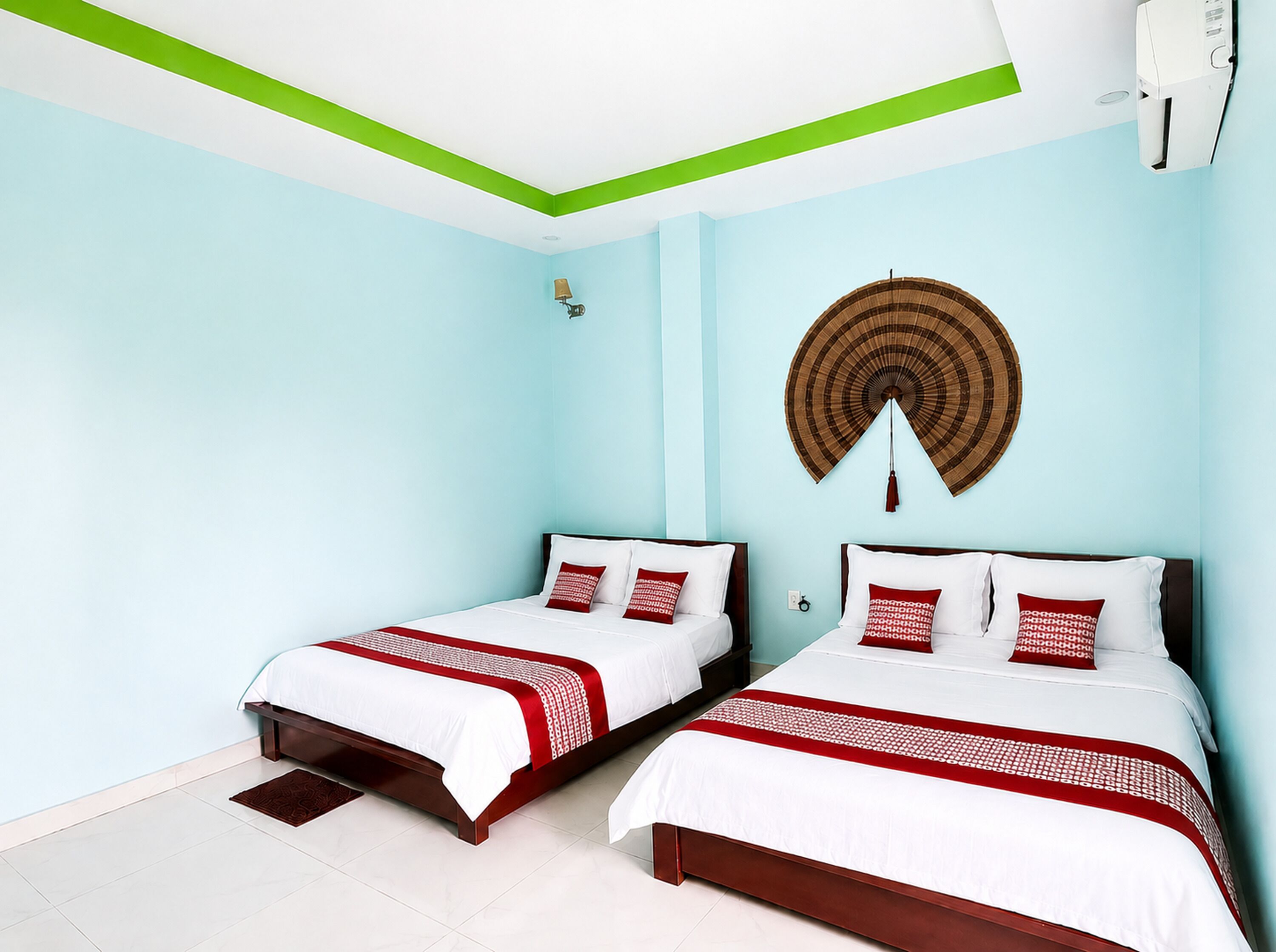 Ein Hotelzimmer mit zwei Betten, hellblauen Wänden, einem mit grünem Zierstreifen versehenen Decke, einem Wandventilator und einem braunen gewebten Wandbehang.