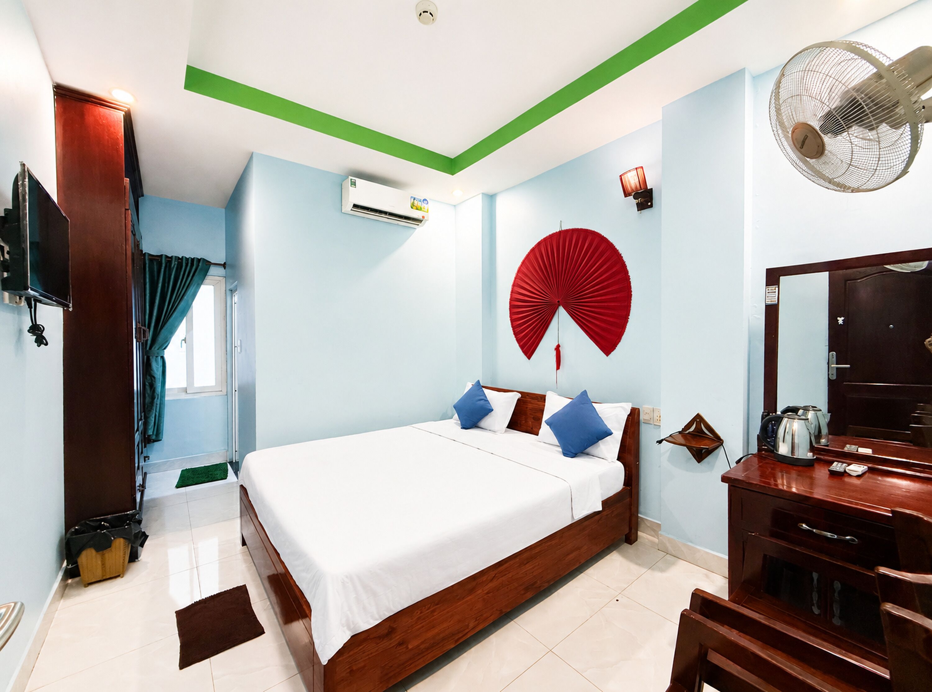 A hotel room with a bed, a fan, a mirror, a dresser, and a window with curtains.