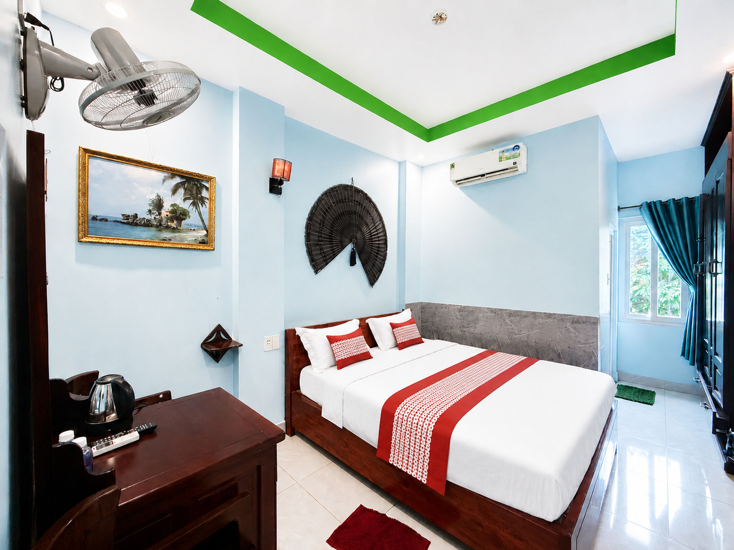 A hotel room with a bed, bedside table, lamp, wall fan, air conditioning unit, and a window with curtains.