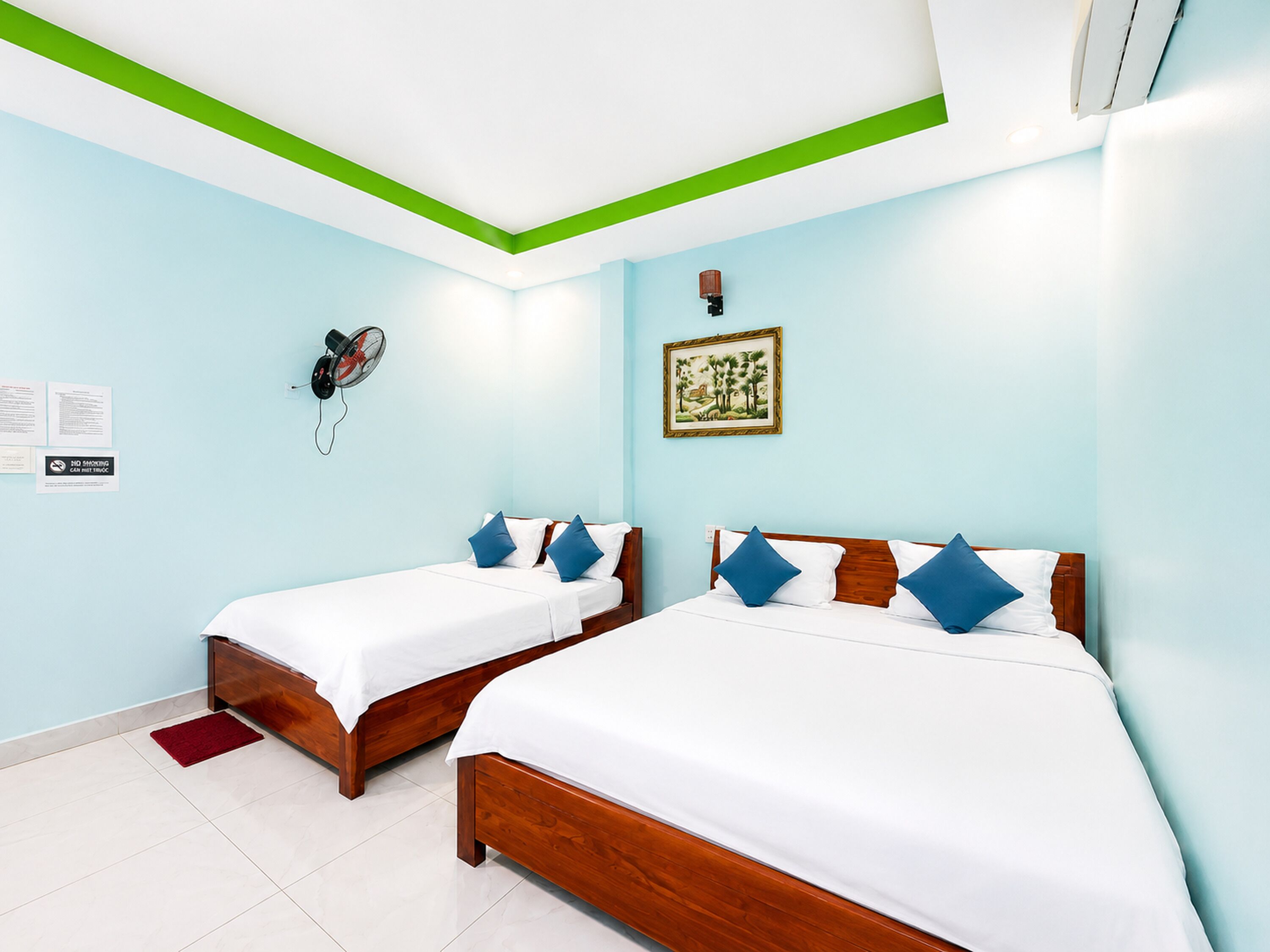 A hotel room with two wooden beds, a ceiling fan, a wall-mounted air conditioner, and a framed picture.