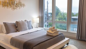 Een net opgemaakt bed met kussens, een bedlampje en uitzicht op de omgeving door een groot raam.