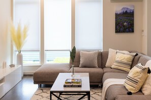 Un salon avec un canapé d’angle, une table basse avec des livres, un vase contenant des plumes d’herbe des pampas et un tableau accroché au mur.