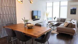 Een moderne woonkamer met een houten eettafel, grijze stoelen, een bank, een televisie en een groot raam.