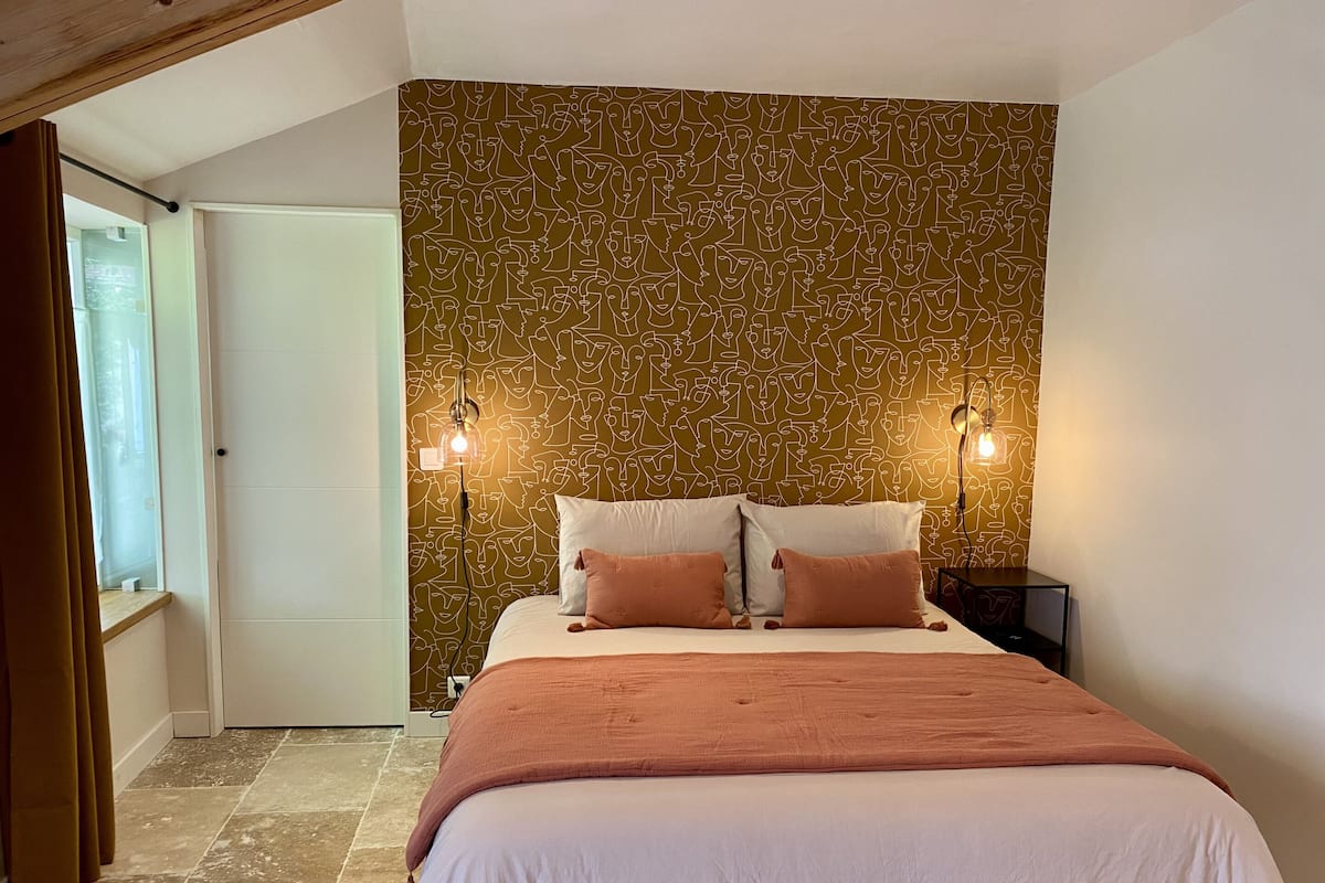A bedroom with a patterned wallpaper, a bed with two pillows, a nightstand, and two wall-mounted lamps.