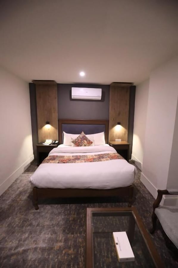 A hotel room with a bed, bedside lamps, a nightstand, a framed picture on the wall, and an air conditioning unit.