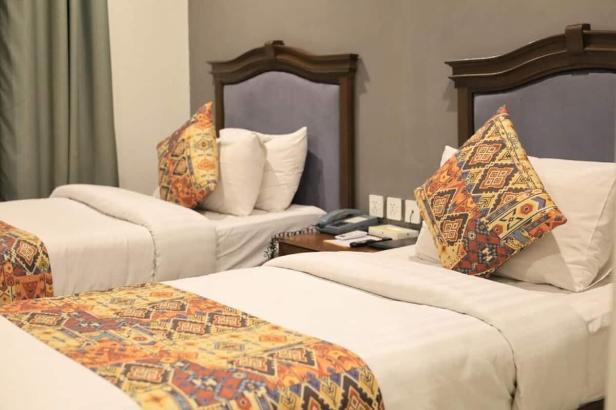 Una habitación de hotel con dos camas, cada una con una colcha y cojines estampados, una mesita de noche con teléfono y un enchufe en la pared.