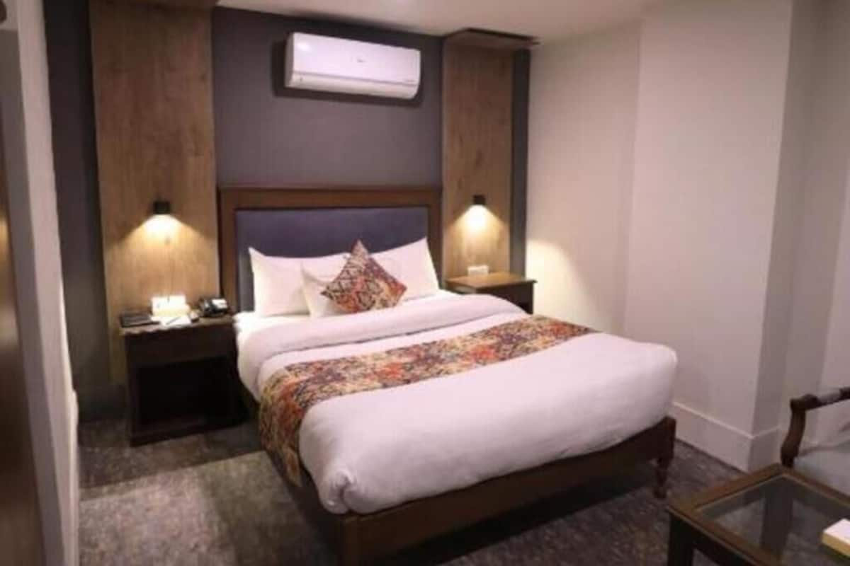 Habitación de hotel con cama, dos mesitas de noche, un buró, unidad de aire acondicionado y una pequeña mesa con una silla.